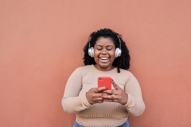 Chica africana con curvas escuchando música de la lista de reproducción mientras mira en el teléfono móvil - Chica milenaria divirtiéndose con las tendencias tecnológicas - Enfoque en la cara