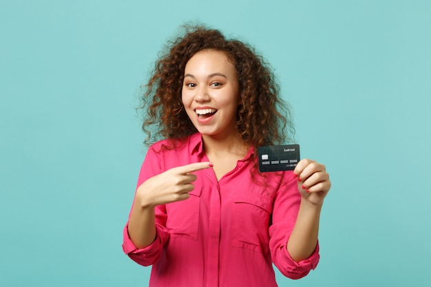 Chica africana emocionada en ropa casual rosa apunta el dedo índice en la tarjeta bancaria de crédito aislada sobre fondo de pared azul turquesa en estudio. Personas sinceras emociones, concepto de estilo de vida. Simulacros de espacio de copia.