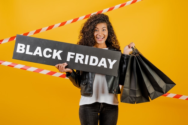 Foto chica africana emocionada con signo de viernes negro y bolsas de papel aisladas sobre amarillo con cinta de señal