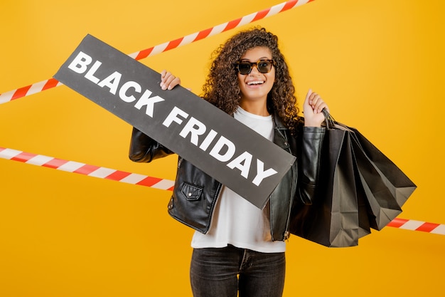 Foto chica africana emocionada con signo de viernes negro y bolsas de papel aisladas sobre amarillo con cinta de señal