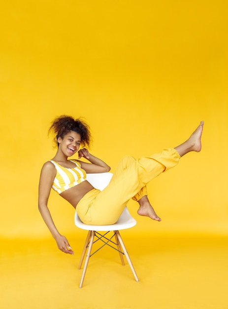Una chica africana feliz usa una elegante tela amarilla se sienta en una silla aislada en el fondo