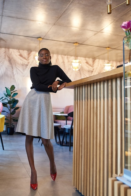 Chica africana de moda sentado en la mesa de café disfrutando de la bebida de capuchino por la mañana joven negra