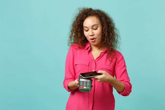 Chica africana sorprendida mantenga terminal de pago de banco moderno inalámbrico de teléfono móvil para procesar adquirir pago con tarjeta de crédito aislado sobre fondo azul turquesa. Concepto de estilo de vida de personas. Simulacros de espacio de copia.