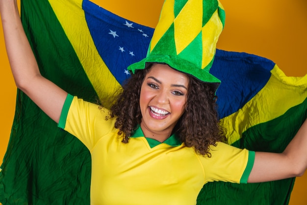 Chica afro animando al equipo brasileño favorito, sosteniendo la bandera nacional en fondo amarillo.