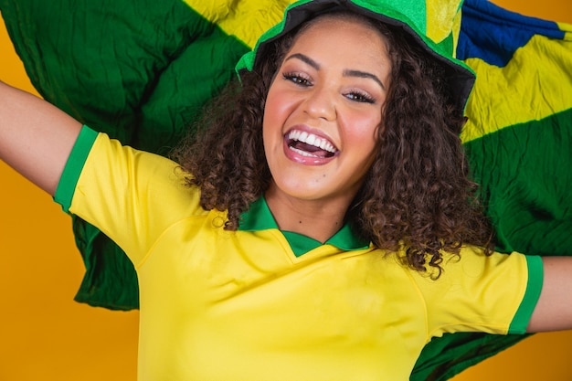 Chica afro animando al equipo brasileño favorito, sosteniendo la bandera nacional en fondo amarillo.