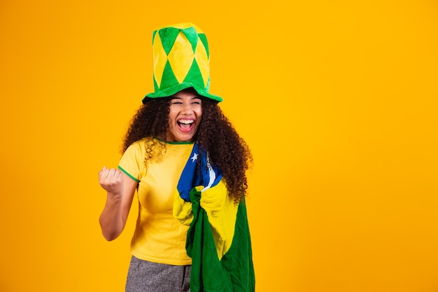 Chica afro animando al equipo brasileño favorito, sosteniendo la bandera nacional en fondo amarillo.