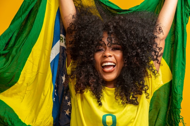 Chica afro animando a su equipo brasileño favorito, sosteniendo la bandera nacional en la pared amarilla.