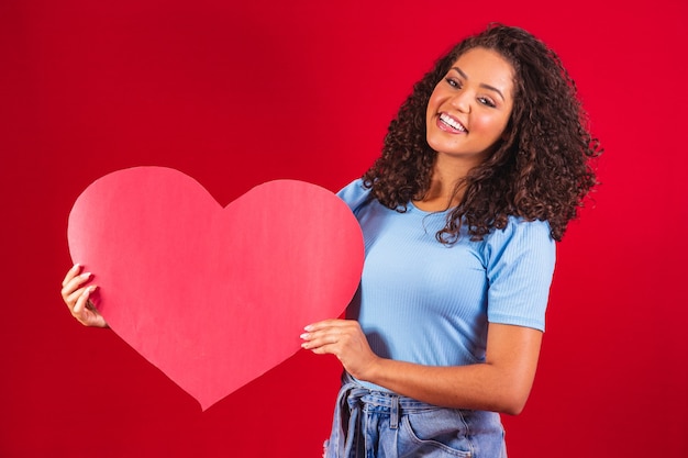 Chica afro sosteniendo en las manos amour en forma de corazón aislado sobre fondo rojo.