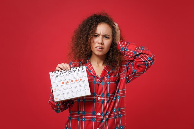 Chica afroamericana cansada en pijama ropa de casa descansa en casa aislada sobre fondo rojo. Relájese el concepto de estilo de vida de buen humor Sostenga el calendario de períodos femeninos para verificar los días de menstruación ponga la mano en la cabeza.