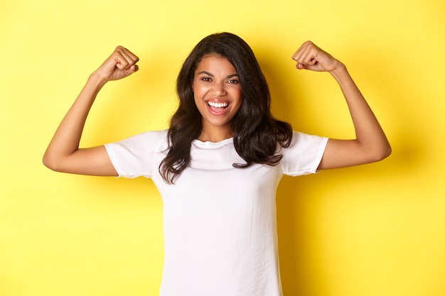 Chica afroamericana confiada y alegre flexionando bíceps y sonriendo con un aspecto fuerte y asertivo