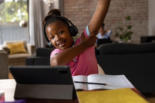 Una chica afroamericana feliz con auriculares levantando la mano durante la lección en línea en casa