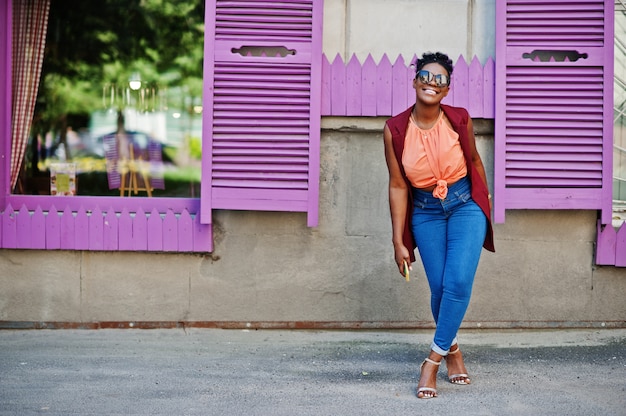 Chica afroamericana en gafas de sol presentado contra ventanas de color púrpura con teléfono celular a mano.