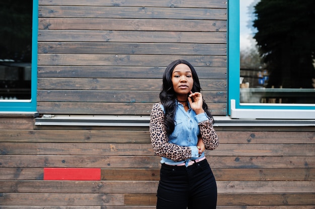 Chica afroamericana hipster con camisa de jeans con mangas de leopardo posando en la calle contra la casa de madera con ventanas.