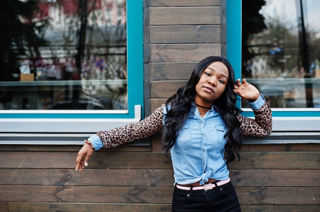 Chica afroamericana hipster con camisa de jeans con mangas de leopardo posando en la calle contra la casa de madera con ventanas.