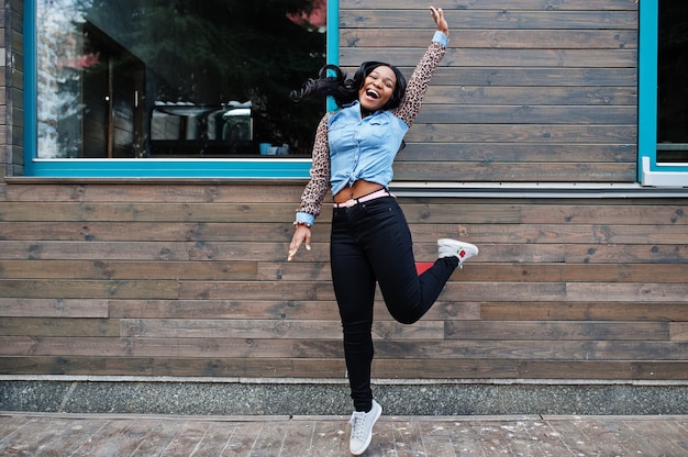 Chica afroamericana hipster con camisa de jeans con mangas de leopardo salta en la calle contra la casa de madera con ventanas.