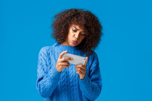 Chica afroamericana linda centrada seria que juega un juego interesante en el teléfono inteligente girando el teléfono móvil, sosteniendo el teléfono horizontalmente, haciendo pucheros perplejos, de pie azul