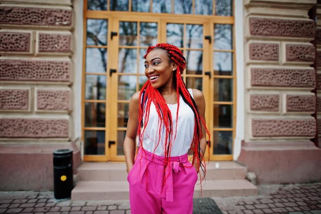 Chica afroamericana de moda en pantalones rosas y rastas rojas posadas al aire libre