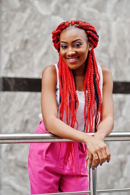 Chica afroamericana de moda en pantalones rosas y rastas rojas posadas al aire libre