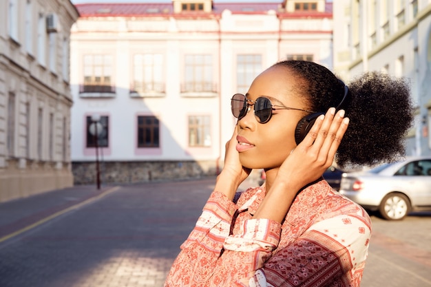 Chica afroamericana negra tiene un paseo por la ciudad de noche y escucha música en auriculares. Chica con gafas de sol en verano sonríe y mirando el atardecer.