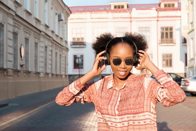 Chica afroamericana negra tiene un paseo por la ciudad de noche y escucha música en auriculares. Chica con gafas de sol en verano sonríe y mirando el atardecer.