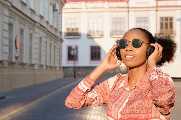 Chica afroamericana negra tiene un paseo por la ciudad de noche y escucha música en auriculares. Chica con gafas de sol en verano sonríe y mirando el atardecer.