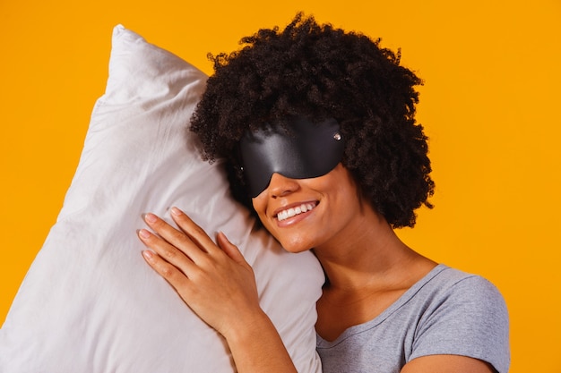Chica afroamericana en pijama y antifaz para dormir está abrazando una almohada y sonriendo, sobre un fondo amarillo