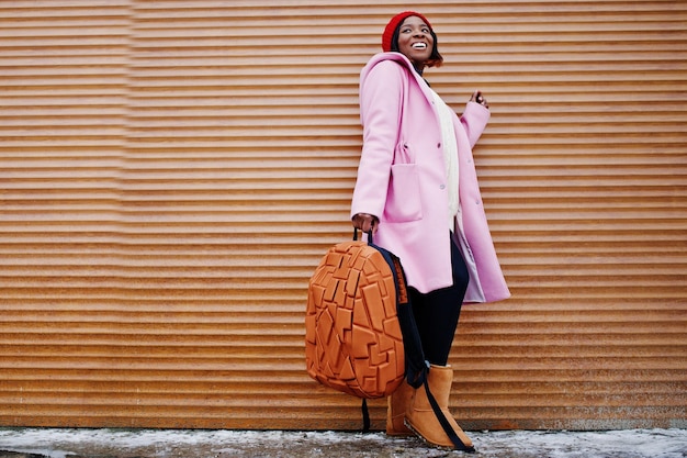 Chica afroamericana con sombrero rojo y abrigo rosa con mochila contra persianas naranjas