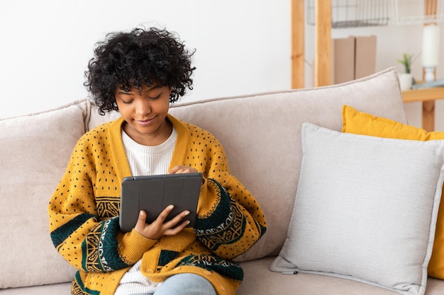 Chica afroamericana sosteniendo una tableta digital con pantalla táctil escribiendo una página de desplazamiento en casa Mujer con un dispositivo de tecnología móvil navegando por Internet usando aplicaciones de redes sociales jugando juegos Estudiar trabajo de compras en línea