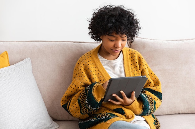 Chica afroamericana sosteniendo una tableta digital con pantalla táctil escribiendo una página de desplazamiento en casa Mujer con un dispositivo de tecnología móvil navegando por Internet usando aplicaciones de redes sociales jugando juegos Estudiar trabajo de compras en línea