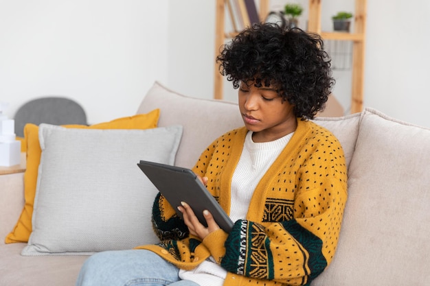 Chica afroamericana sosteniendo una tableta digital con pantalla táctil escribiendo una página de desplazamiento en casa Mujer con un dispositivo de tecnología móvil navegando por Internet usando aplicaciones de redes sociales jugando juegos Estudiar trabajo de compras en línea