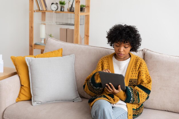 Chica afroamericana sosteniendo tableta digital pantalla táctil escribiendo página de desplazamiento en casa mujer con mobi