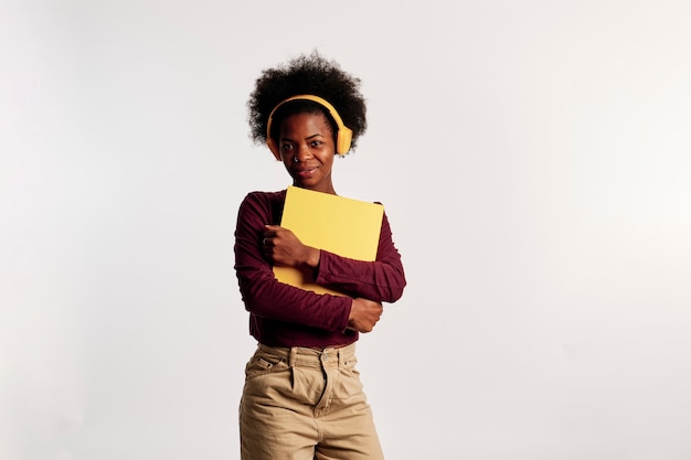 Chica afroamericana en suéter marrón posa con su carpeta amarilla mientras escucha música.