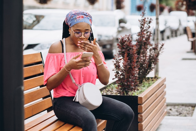 Chica afroamericana usando su teléfono móvil mientras está sentado en un banco de madera