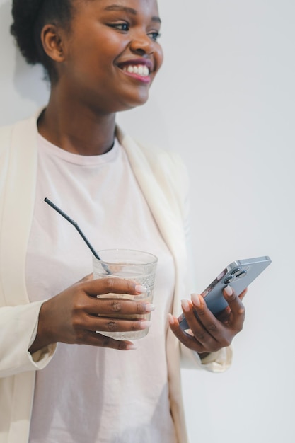Chica afroamericana usando un teléfono inteligente para enviar mensajes mientras bebe un café helado en un café sonriendo mirando hacia otro lado Mujer relajándose en vacaciones Conectado a internet inalámbrico