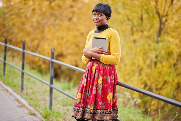 Chica afroamericana en vestido amarillo y rojo en el parque de otoño dorado con tableta en las manos