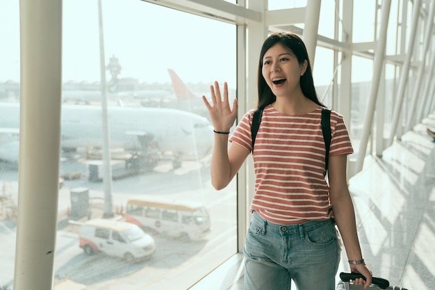 Chica agita su mano al llegar al aeropuerto de hong kong sosteniendo una maleta y sonríe. joven mujer china asiática caminando en el pasillo a lo largo de la ventana fuera de la puerta diciendo hola para recoger a su familia y amigos ch