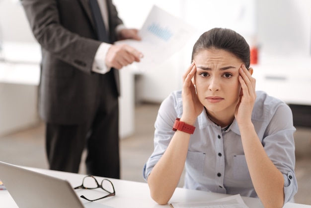 Foto chica agotada sentada en su lugar de trabajo con la cabeza apoyada en las manos presionando los labios mientras mira al frente