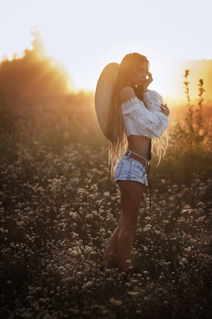 Chica al atardecer en un sombrero. campo de flores blancas. rastas. hermoso look de verano