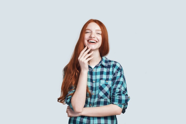 Chica alegre, alegre y despreocupada con un hermoso cabello largo rojo riendo mientras está parado sobre fondo gris