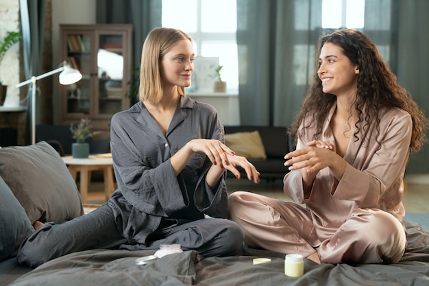 Chica alegre aplicando producto cosmético natural hecho a mano mientras está sentada en la cama por la mañana, su amiga con smartphone disparándole en la cámara