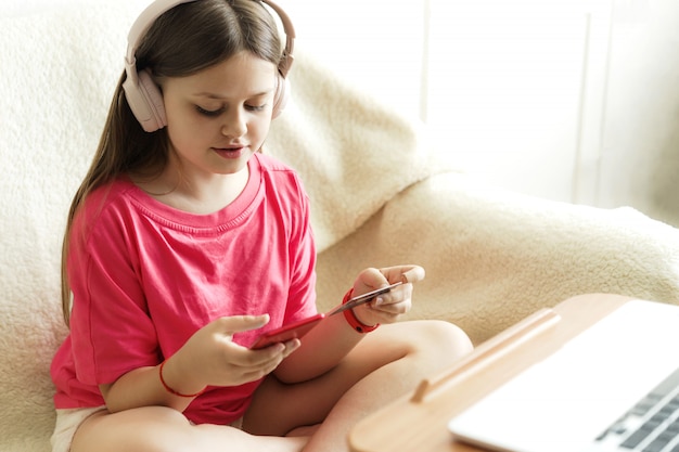 Chica alegre en auriculares y una camiseta rosa se sienta con un teléfono en la mano y una tarjeta de crédito