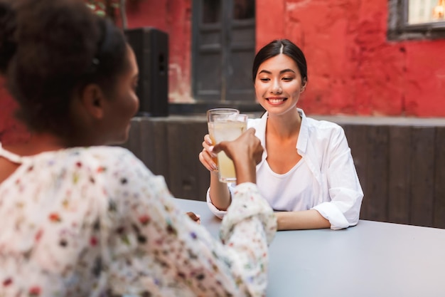 Chica alegre con camisa blanca bebiendo cócteles con un amigo mientras pasa felizmente el tiempo en el patio de la cafetería