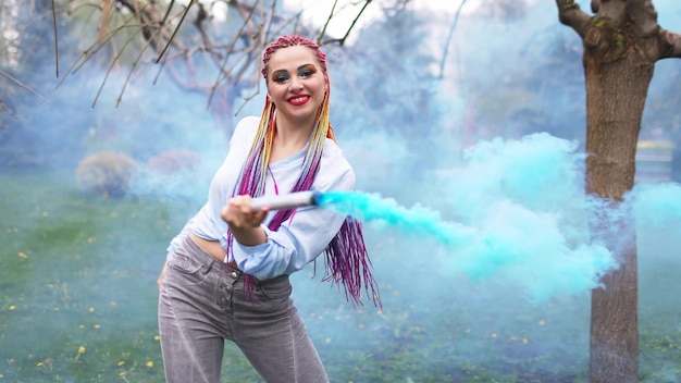 Una chica alegre con una camisa y jeans con trenzas de arco iris brillantes y maquillaje inusual. Ella baila escondida en un espeso humo artificial azul