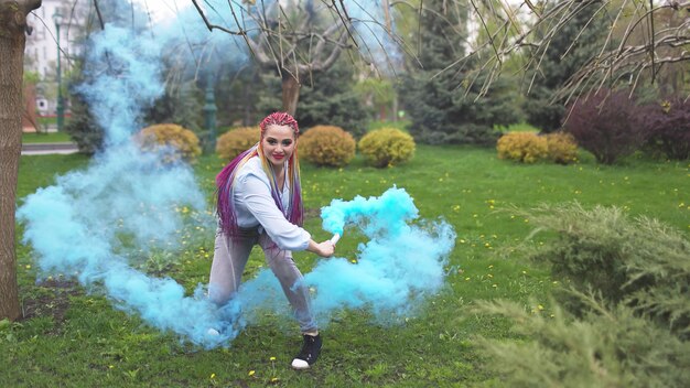 Una chica alegre con una camisa y jeans con trenzas de arco iris brillantes y maquillaje inusual. Ella baila escondida en un espeso humo azul artificial con el telón de fondo del parque de primavera