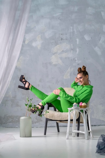 Chica alegre en camisa verde y pantalones se sienta en una silla cerca de la mesa con café y pastelitos Hermosa mujer en estudio gris