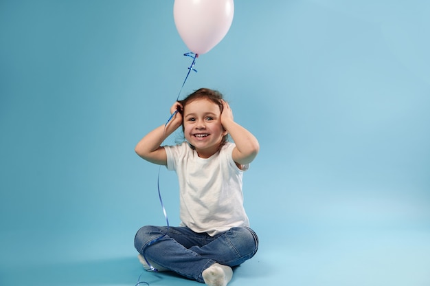 Foto chica alegre en camiseta blanca y jeans azul se sienta en la superficie azul con un globo rosa en la mano