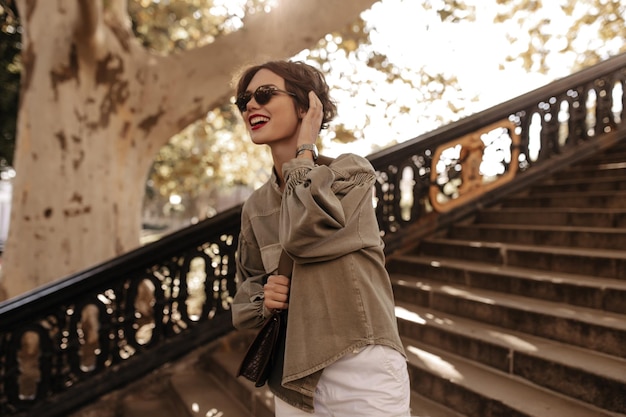 Chica alegre con chaqueta de mezclilla verde oliva y pantalones blancos sonriendo en el fondo de las escaleras Señora alegre con bolso y gafas de sol posa afuera