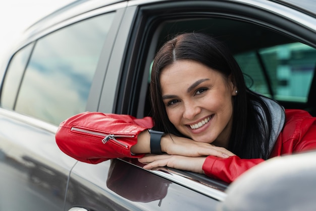 Chica alegre conduce su auto nuevo con placer Ella sonríe con placer y mira la cámara Concepto de pasajero