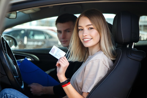 Chica alegre conduciendo un coche de entrenamiento con una tarjeta de licencia de conducir en sus manos.