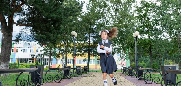 Chica alegre y divertida con sonrisa desdentada en uniforme escolar con lazos blancos corriendo en el patio de la escuela Regreso a la escuela 1 de septiembre Alumno feliz con mochila Educación primaria movimiento de clase primaria